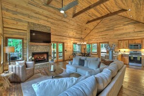 A log cabin interior with a stone fireplace, a large sofa, and a kitchen area. - Luxury Blue Ridge 3BR Cabin w/ Outdoor Living Area & Hot Tub (Epworth)