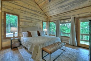 A wooden cabin bedroom with a large bed, a bench, and multiple windows offering a forest view. - Luxury Blue Ridge 3BR Cabin w/ Outdoor Living Area & Hot Tub (Epworth)