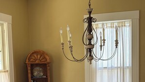 A dining room with a round wooden table, six black chairs, a chandelier, a bookshelf, and a window with sheer curtains.