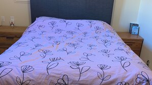 Una cama con una colcha de motivos florales, un cabecero oscuro, una mesita de noche con libros y un enchufe en la pared.