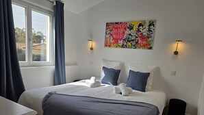 A hotel room with a bed, a desk, a window with curtains, and a colorful artwork on the wall. - Les Photinias Giens Peninsula Beach King-Size Bed (Hyères)