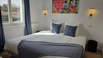 Un dormitorio con cama, dos almohadas, una colcha, un cuadro en la pared y una ventana con cortinas.