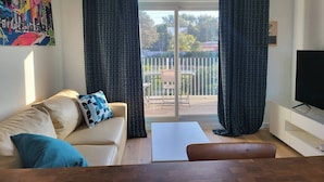 A living room with a sofa, a TV, and a balcony with curtains. - Les Photinias Giens Peninsula Beach King-Size Bed (Hyères)