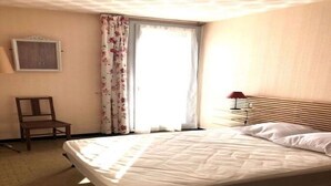 Ein Hotelzimmer mit Bett, Sessel, Fenster mit Vorhängen und Nachttisch mit Lampe.