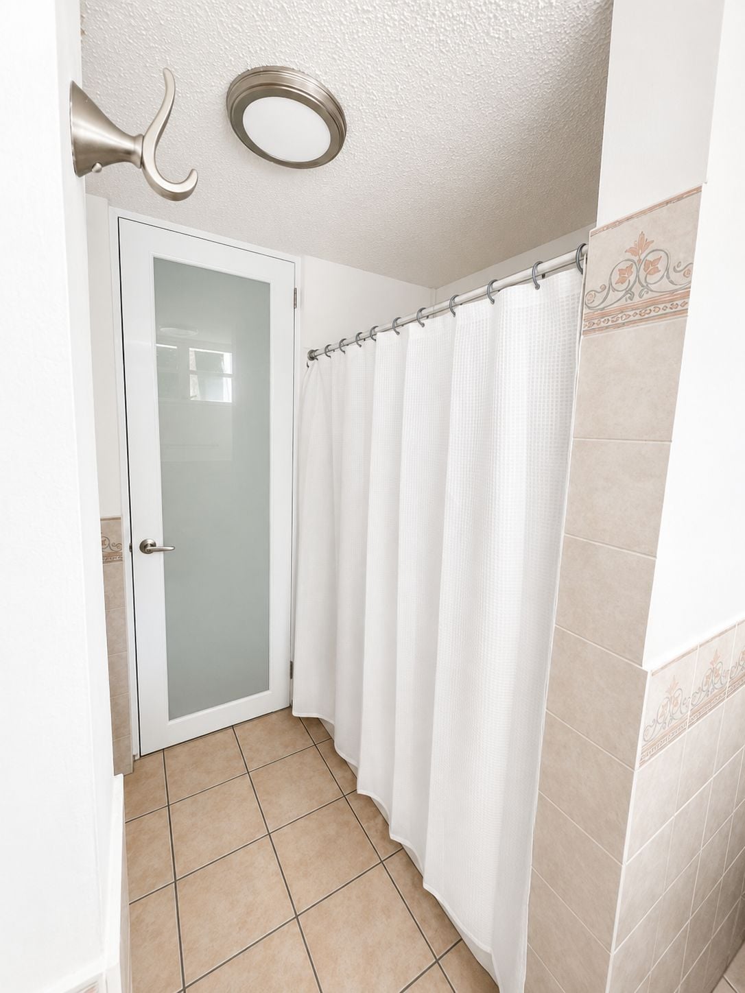 Un baño con puerta de cristal, cortina de ducha blanca y suelo de baldosas color beige.