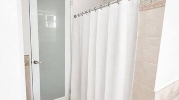 Un baño con puerta de cristal, cortina de ducha blanca y suelo de baldosas color beige.