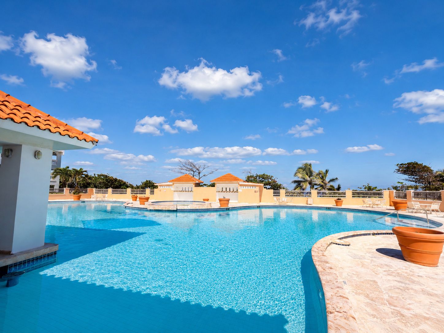 A clear blue pool with a tiled deck, surrounded by buildings with orange roofs and a clear sky.