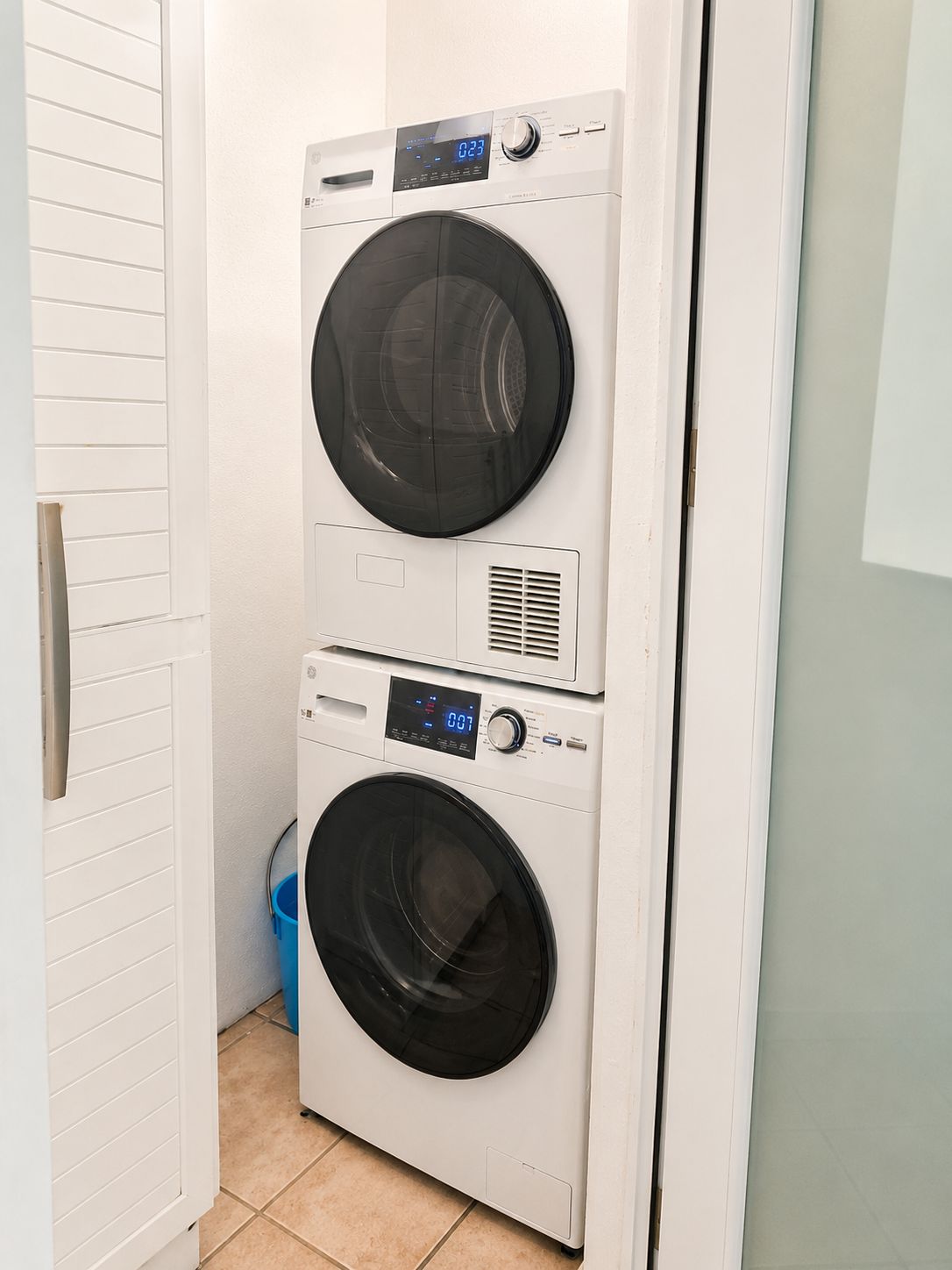 Une machine à laver et un sèche-linge superposés dans une buanderie, avec une porte blanche et un sol carrelé.