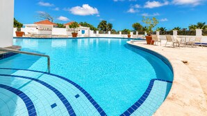 Una piscina azul cristalina con bordes de azulejos, rodeada de sillas de descanso y plantas en macetas, bajo un cielo azul brillante.