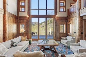 A spacious room with a large window, wooden walls, a sofa, a coffee table, and a view of a mountainous landscape. - Secluded 160-Acre Jackson Hole Ranch and Mountain Views (Jackson)