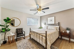 Une chambre à coucher comprenant un lit, un ventilateur de plafond, une chaise, une petite table, un miroir, une fenêtre et une plante.