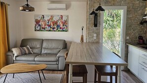 A modern living room with a grey sofa, a wooden dining table, and a kitchen area with a stone wall and a window.