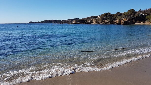 A beach with clear blue water, a sandy shore, and buildings on a rocky hillside in the background.