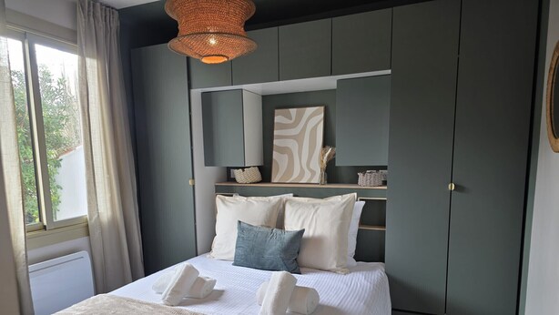 A bedroom with a bed, a bedside table, a hanging light fixture, a window with curtains, and a wall-mounted shelf with decorative items. - - (Hyères)