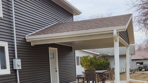 A covered patio area with a table and chairs, a small tree, and a house with dark siding and a white door. - Spacious 4-bedroom house in Cherokee with AC (Cherokee)