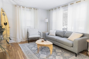 A living room with a grey sofa, a wooden coffee table, a white armchair, a surfboard, and a patterned rug.