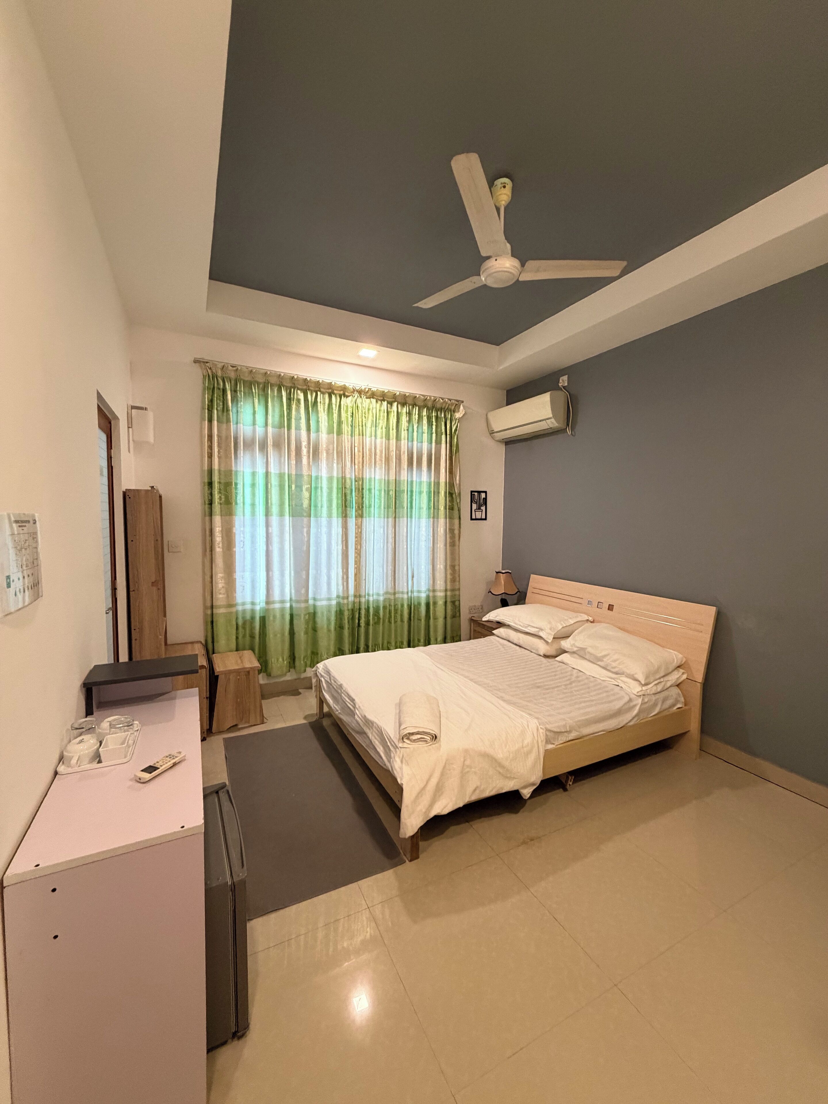 A neatly arranged hotel room with a bed, a desk, a chair, a ceiling fan, and an air conditioning unit.