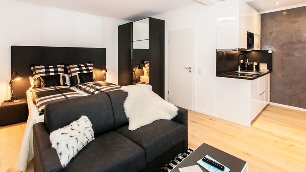 A modern bedroom with a large bed, a sofa, a mirror, and a small kitchenette. - Exclusive studio business apartment (Neu-Isenburg)