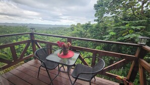 A wooden deck with two chairs and a table, overlooking a lush green landscape. - Mi Verde Horizonte "Panoramic view in Utuado | Nearby river + Nature + WiFi". (Utuado)