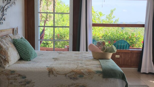 A bedroom with a bed, a ceiling fan, a window with curtains, and a view of greenery outside. - Mi Verde Horizonte "Panoramic view in Utuado | Nearby river + Nature + WiFi". (Utuado)