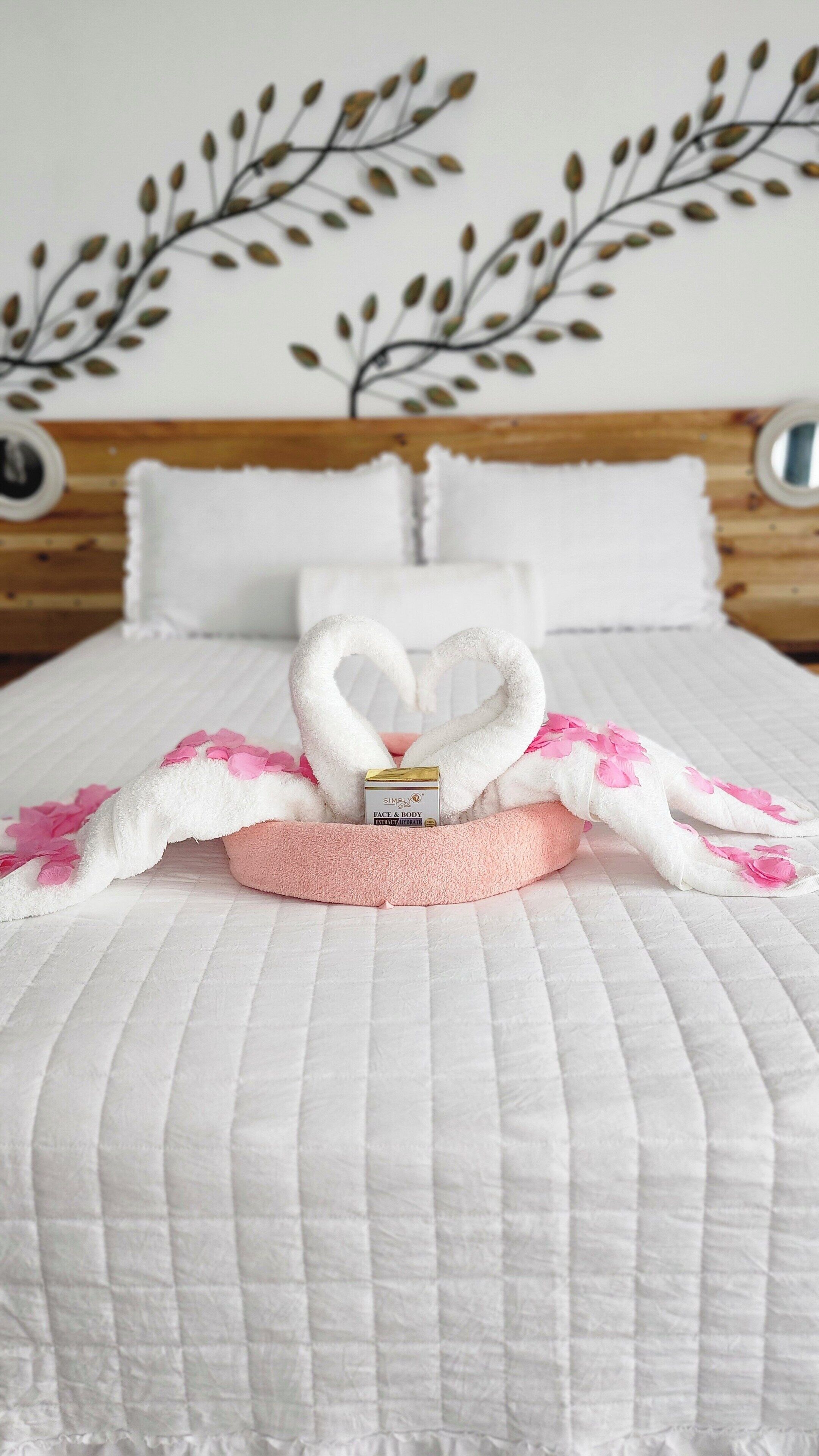A neatly made bed with a heart-shaped towel arrangement, a pink pillow, and a bottle of facial cream.