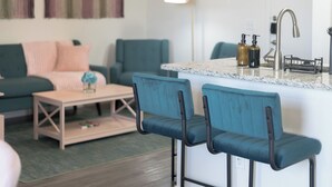 Een moderne woonkamer met een blauwgroene bank, een salontafel, en een keuken met een marmeren aanrecht en barkrukken.