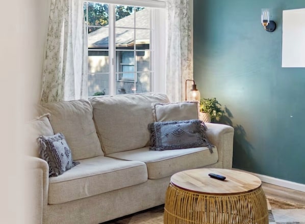 A beige sofa with two pillows, a round wicker side table, and a lamp against a teal wall. - Cozy Browne’s Addition Apt Near Downtown (Spokane)