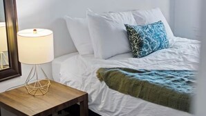 Ein ordentlich bezogenes Bett mit weißen Leinentüchern und einem blau gemusterten Kissen, ein Nachttisch aus Holz mit einer geometrischen Lampe und ein gerahmtes Kunstwerk an der Wand.