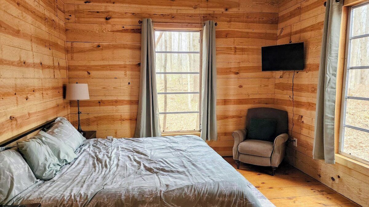 A wooden cabin room with a bed, a chair, a lamp, and a TV mounted on the wall.