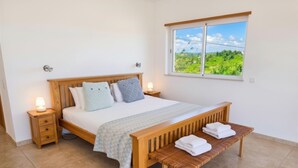 A bedroom with a wooden bed, two bedside tables with lamps, a bench with towels, and a window with a view of greenery. - Casa da Madrugada - Peaceful haven with pool and ocean view in Salema (Salema)
