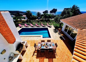 A pool with a view of the sea, a dining area with a table set, and a building with a tiled roof. - Casa da Madrugada - Peaceful haven with pool and ocean view in Salema (Salema)