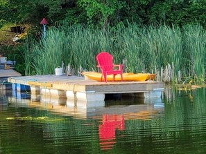 Un muelle con una silla roja y un barco amarillo, rodeado de hierba alta y árboles.