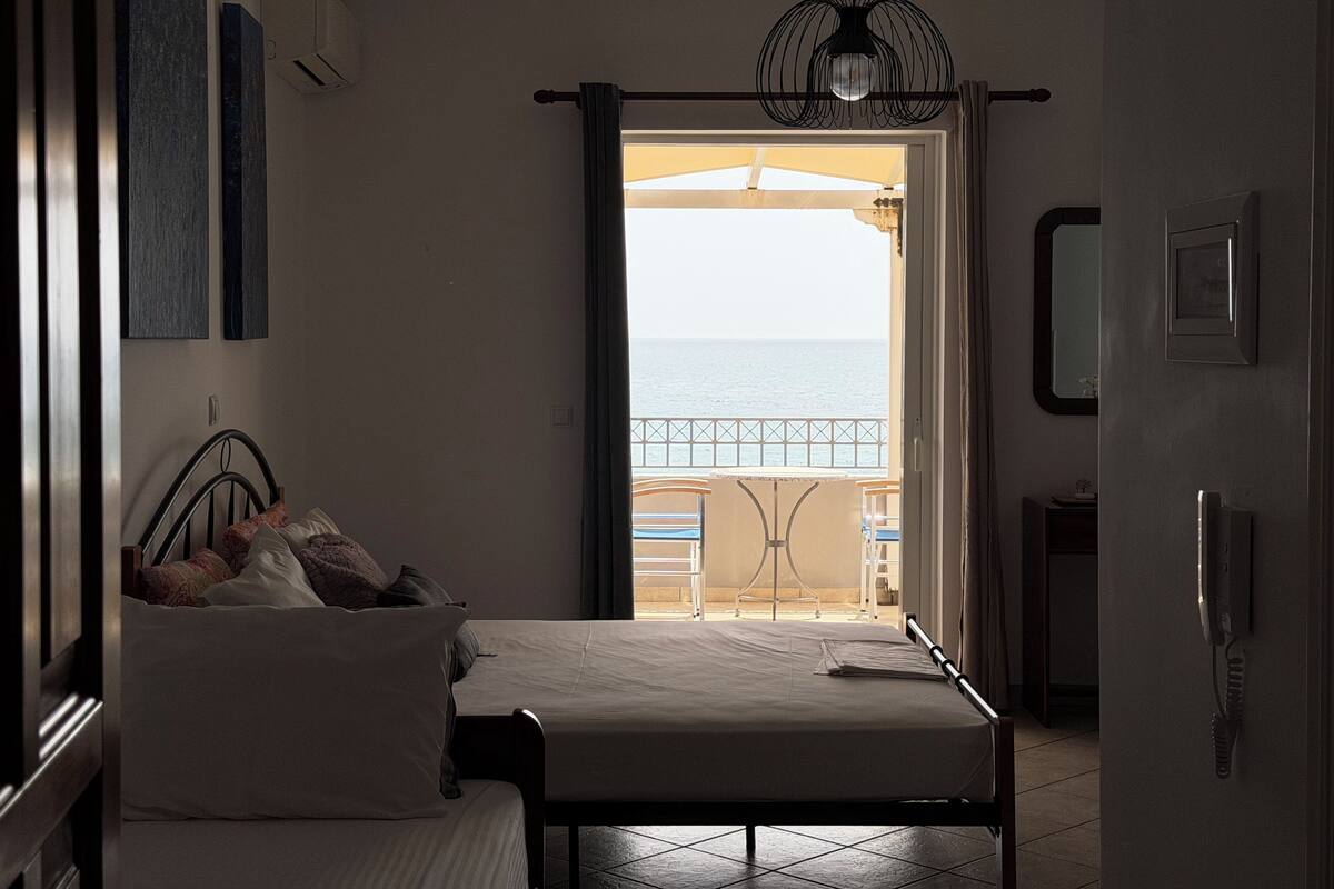 A bedroom with a bed, a bedside table, a hanging light fixture, a door with a phone, and a view of the sea through an open door.