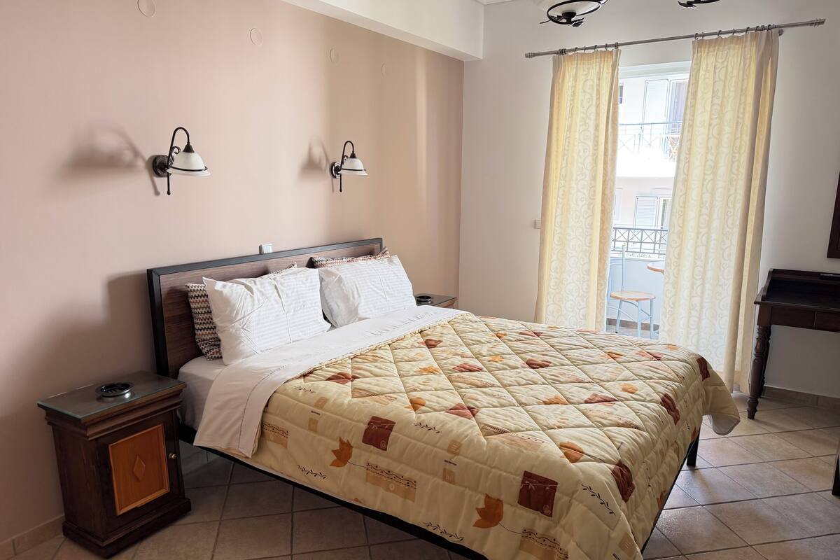 A neatly made bed with a quilted cover, a bedside table with a lamp, and a view of the balcony through curtains.