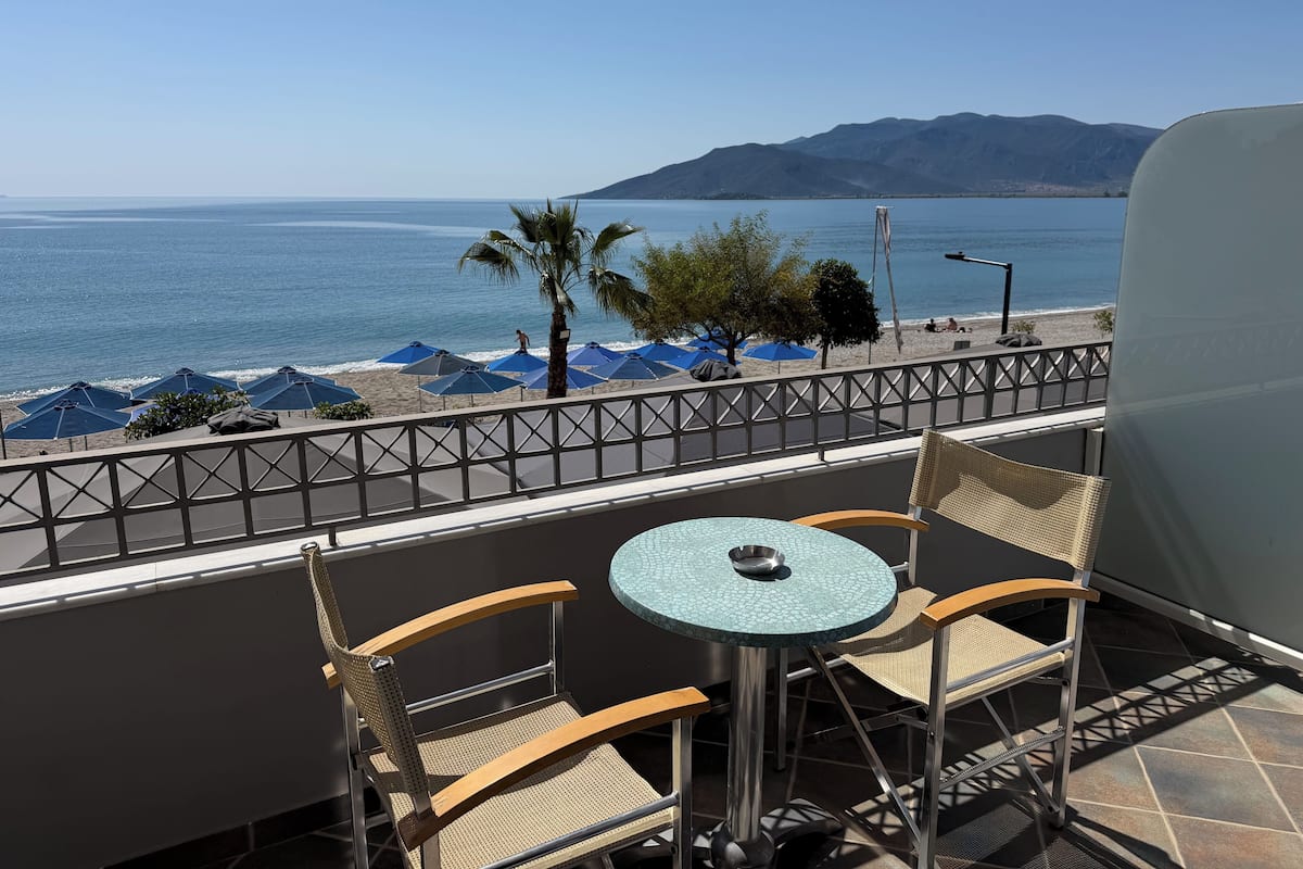 A balcony with a table and chairs, overlooking a beach with umbrellas and a mountain in the distance.