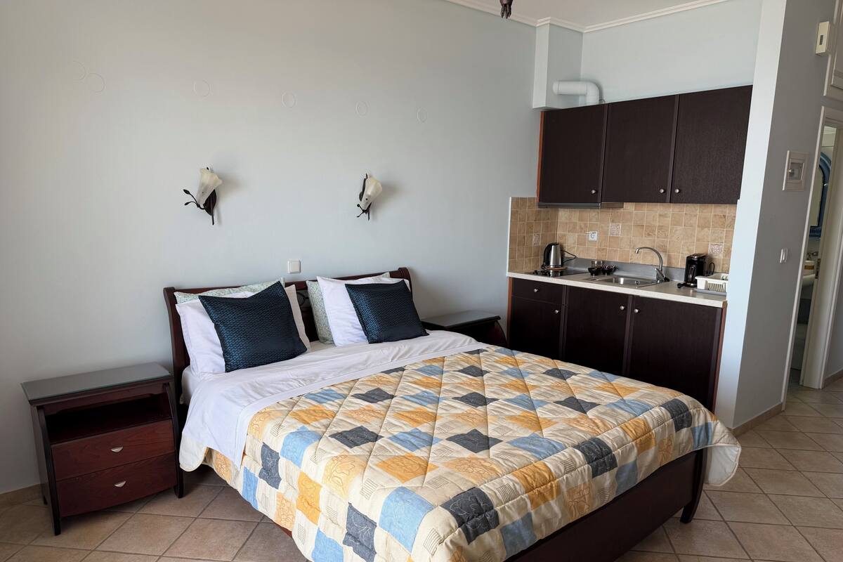 A hotel room with a bed, a nightstand, a small desk, a kitchenette, and a bathroom visible through an open door.