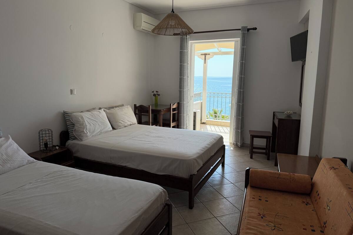A bedroom with two beds, a sofa, a table, a TV, and a balcony with a view of the sea.