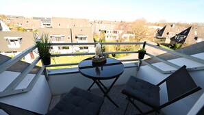 Un piccolo balcone con due sedie, un tavolo, piante in vaso e vista su edifici residenziali.