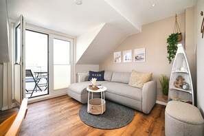 A cozy living room with a grey sofa, a small round table, and a grey rug. There is a window with blinds, a plant, and a shelf with decorative items. - Hus Willem - Kuschelkoje Vacation Apartment (Büsum)