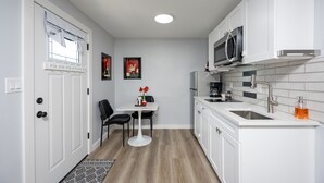 A compact kitchen with white cabinetry, a microwave, and a sink. There is a small dining table with chairs and a rug leading to the door. - Tiny House: 1BR w/ KING Bed, Pool by Raymond James (Tampa)