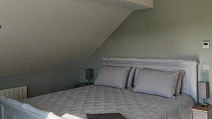Un dormitorio con techo inclinado, una cama con colcha estampada, dos almohadas, una mesita de noche con una lámpara y un termostato fijado a la pared.
