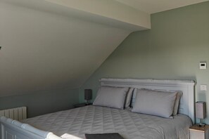 A bedroom with a sloped ceiling, a bed with a patterned bedspread, two pillows, a bedside table with a lamp, and a wall-mounted thermostat. - 4 Bed House Sleeps 8 - Enclosed Garden - Parking (Killarney)