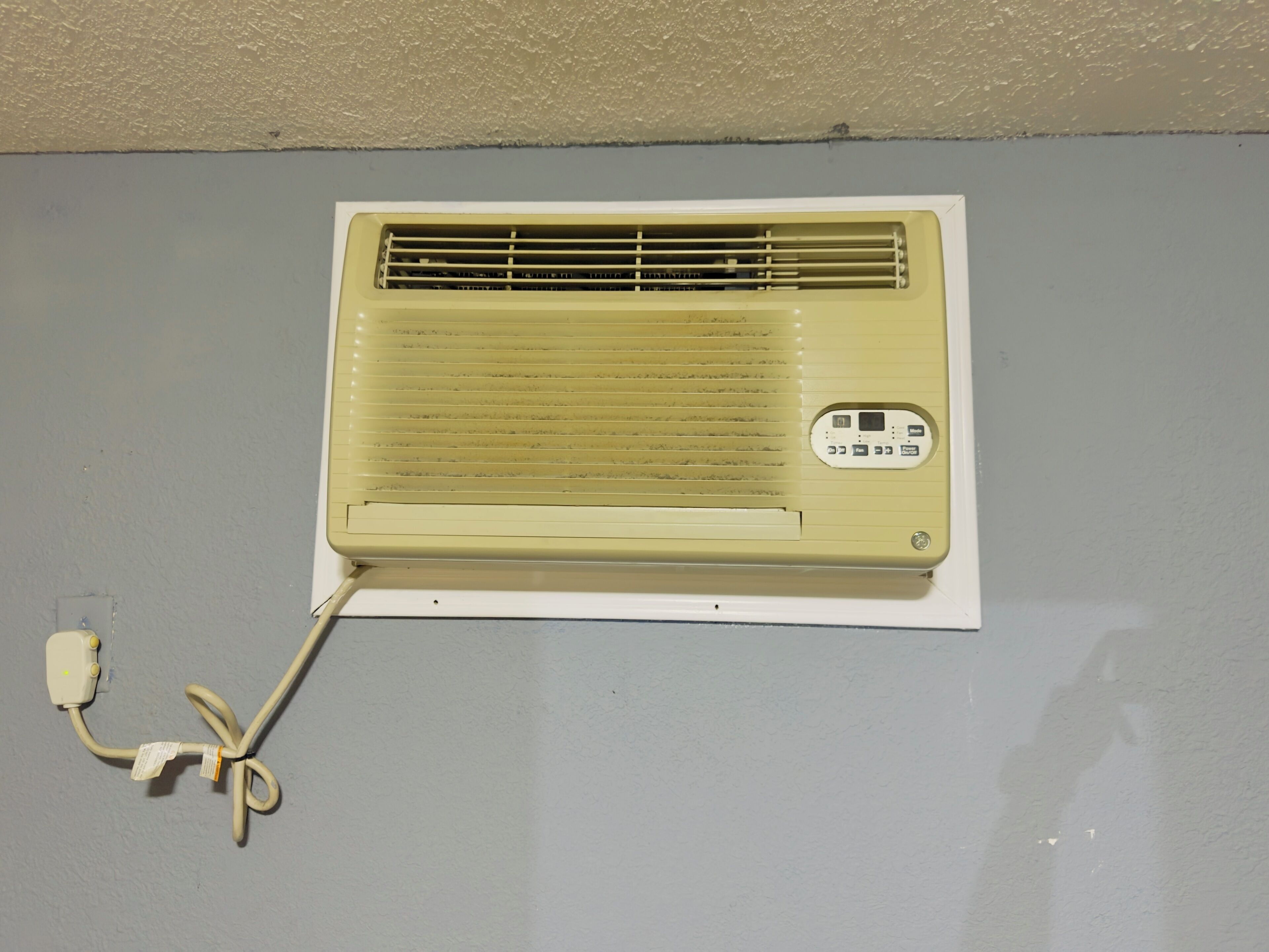 Un climatiseur ancien, doté d’un panneau de commande et relié à une prise électrique murale.