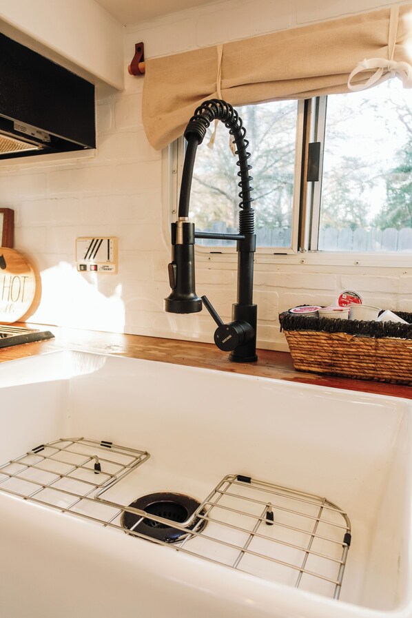 Una cocina con un lavamanos blanco, un grifo negro, una ventana con vistas a árboles, un letrero de madera en la pared y una cesta sobre la encimera.