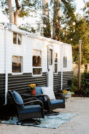 A white RV with a black lower half, a wicker chair with blue cushions, and a small table with a potted plant. - Cozy Glamping Retreat • Sleeps 4 • Family Fun or Romantic Getaway  (Lutz)
