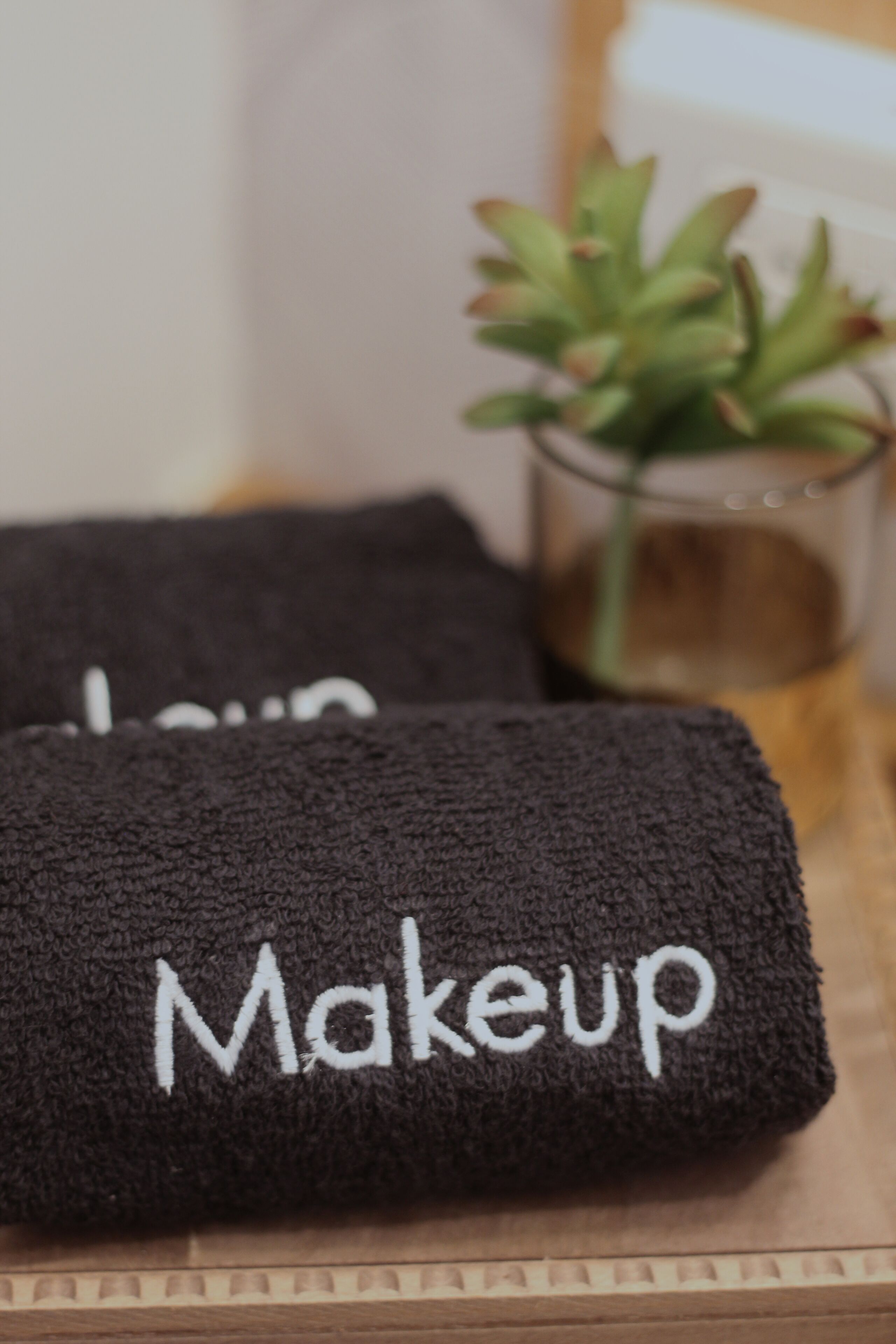 A black towel with the word "Makeup" embroidered on it, placed on a wooden surface.