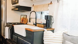 Eine kompakte Küche mit Herd, Mikrowelle und Kaffeemaschine. Ein Spülbecken mit Handtuch und eine kleine Kücheninsel mit Korb sind vorhanden. Der Raum verfügt über ein gemütliches Bett mit Kissen und Decke.
