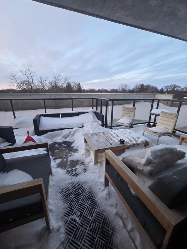 An outdoor seating area with snow-covered furniture and a view of trees and a cloudy sky.