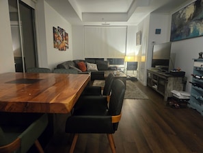 A modern living room with a wooden dining table, black leather chairs, a grey sofa, and a TV.