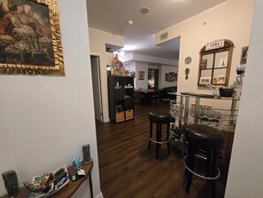 A room with a wooden floor, a dining area with a table and chairs, a bar area with stools and a glass display, and a wall with a framed picture and a mirror.